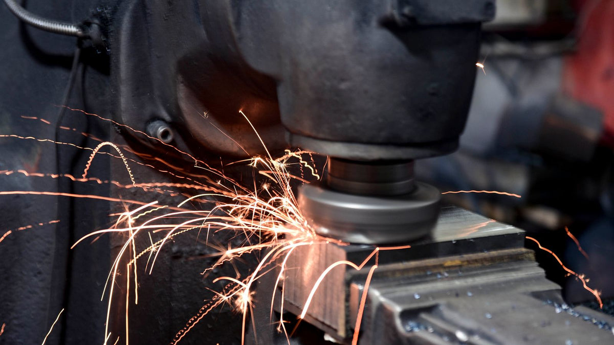 A worker grinding a steel part on a bench grinder with sparks flying off the wheel.