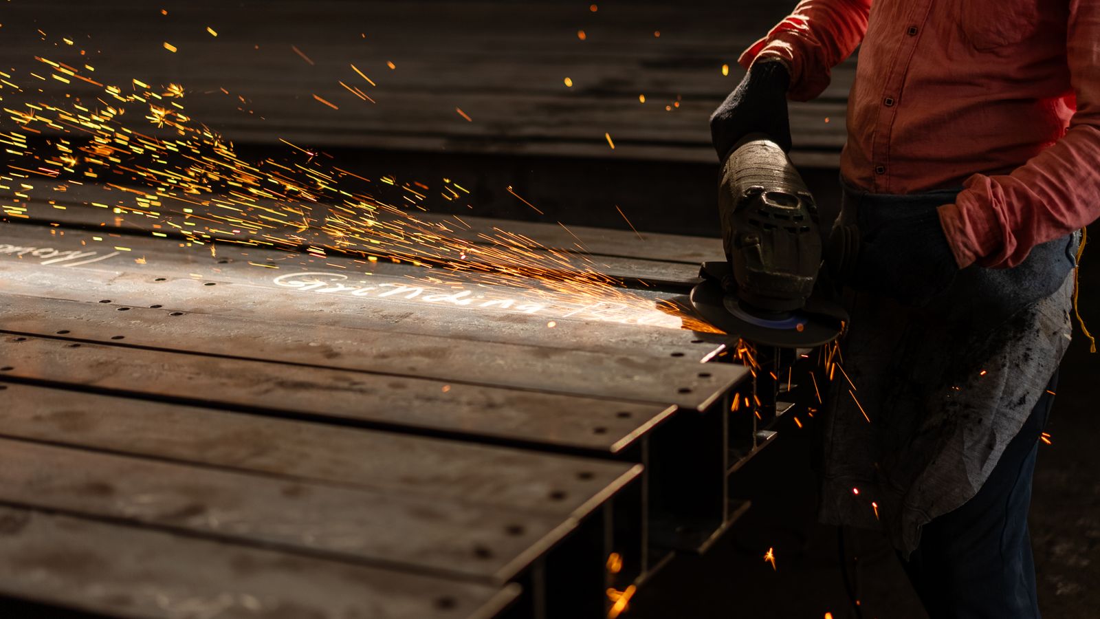 Worker grinding steel beams with an angle grinder, sending sparks across a fabrication shop floor