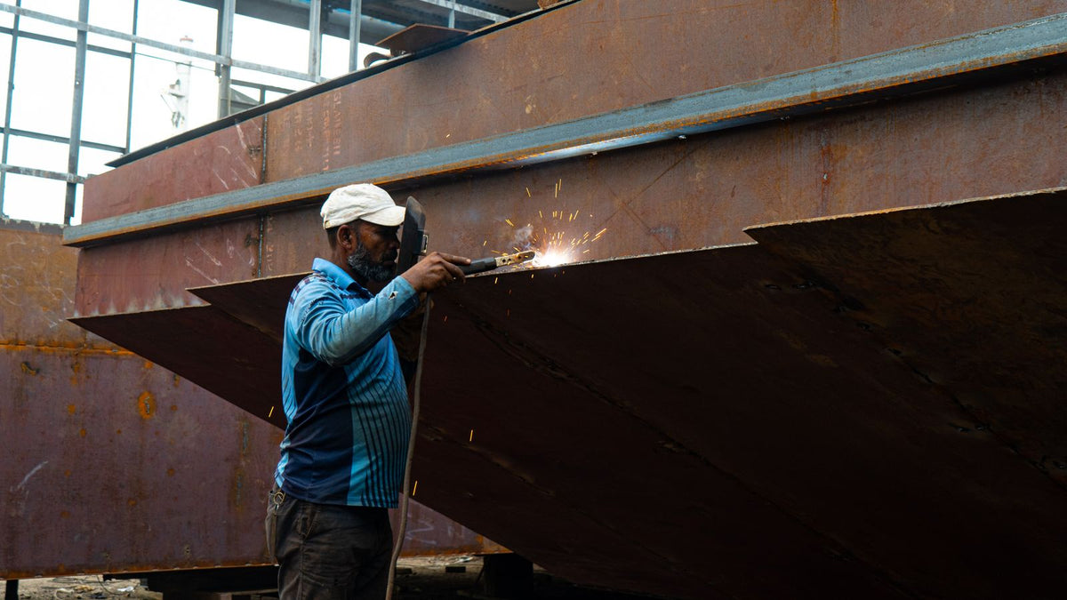 A welder grinding a steel joint on a large ship hull under construction in a shipyard