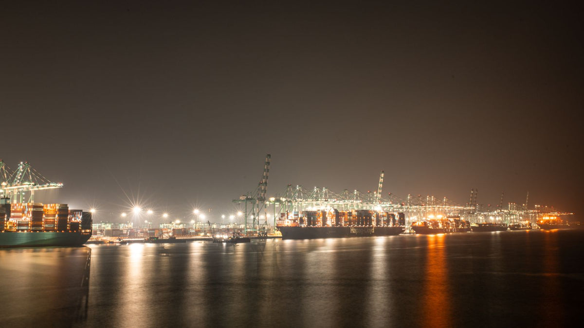 Stacked shipping containers at an industrial port with cranes loading cargo in the background.