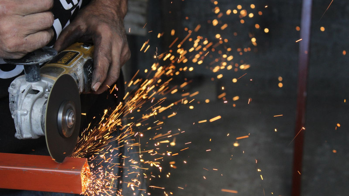 A worker uses an angle grinder with a cutting disc to slice through a steel beam, throwing off sparks.